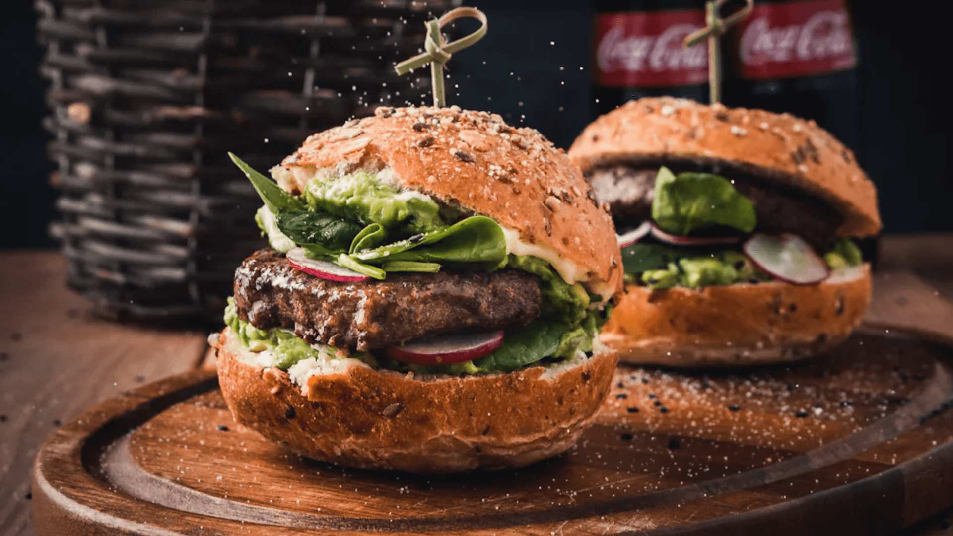Two gourmet beef burgers served on a wooden board, featuring thick patties topped with guacamole, fresh spinach, and sliced radishes on sesame seed buns, with Coca-Cola bottles visible in the background.