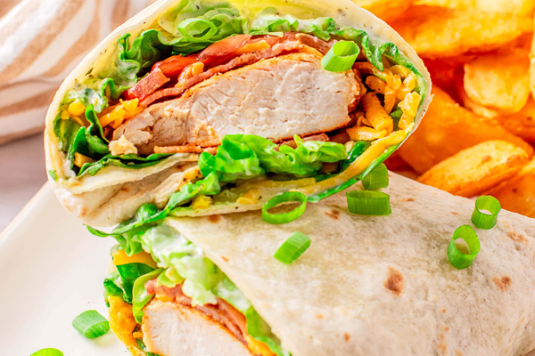 A grilled chicken wrap cut in half and stacked to show the filling of sliced chicken breast, fresh lettuce, tomatoes, and cheese, with seasoned potato wedges in the background.